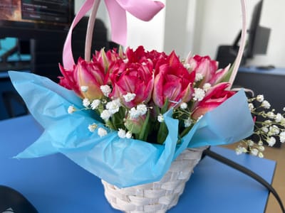 A small, light-colored woven basket containing a bouquet of red and yellow-tipped tulips and white baby's breath rests on a dark blue desk surface. The flowers are wrapped at their base in light blue tissue paper, and the basket's handle is adorned with a pink ribbon tied in a bow. A black computer mouse or similar peripheral is partially visible on the desk to the left, and a black cable extends from the right foreground. The background is blurred, showing an office environment with multiple computer monitors and office chairs. The scene is located in Большевик, Russia.