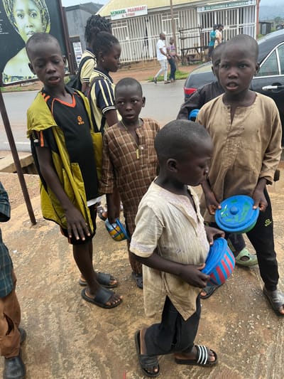 The image captures a group of children on a paved surface in what appears to be an outdoor marketplace or street in Sabojibi, Nigeria. The scene is oriented vertically, with the subjects appearing from bottom to top.

In the foreground, a young boy wearing a yellow and black shirt is looking towards the camera with an open mouth, suggesting he might be speaking or reacting to something. He has dark skin and short, dark hair.

Behind him, a group of children are gathered. Some are seated or crouching. One child, in a light-colored, loosely fitting shirt and dark pants, is holding a blue plastic disc with red and white stripes. Another child, in a brown checkered shirt, is also present. Several children are wearing sandals. In the background, other individuals are visible, and there's a glimpse of buildings and a vehicle.

The overall lighting suggests it is daytime. The children's expressions are difficult to fully discern due to the image's orientation and resolution, but their presence together and the plastic toys suggest a moment of play or casual gathering.

Visible text in the background includes "GLC CLOTHINGS & ACCESSORIES" on a storefront. Another sign reads "AKMAKEL PHARMACY & STORE". The presence of these signs and the general street scene indicates an urban or semi-urban environment.