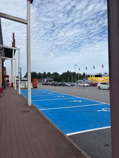 This is a landscape photo taken from a covered walkway, looking out at a parking lot and a building in Pori, Finland. The image is oriented vertically. The foreground shows a red brick pavement and a white metal structure of the walkway. Beyond the walkway is a parking lot with several cars parked. Some of the parking spaces are painted bright blue with white lines and feature a wheelchair symbol, indicating disabled parking. Further back, there is a yellow building with flags and a white car parked in front of it. Trees and a cloudy sky are visible in the background. There is a single orange scissor lift in the parking lot. The time of day appears to be daytime, and the weather is cloudy. There are no people clearly visible in the image. Text visible includes "XXL" on flags and the building, and "XXL SPORT" on a sign on the building. There are also Finnish words on the building and a parking sign with a blue "P".