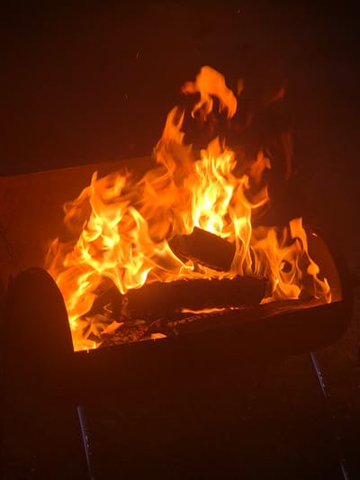 A fire burns in a dark, cylindrical metal fire pit. The flames are predominantly orange and yellow, with some hints of white in the hottest areas. Logs of wood are visible within the fire pit, partially consumed by the flames. The background is dark and indistinct, suggesting an outdoor setting at night. Two small, faint sparks are visible in the upper left quadrant of the image. The fire pit appears to be elevated on short metal legs.
