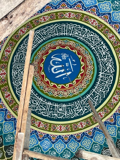 An upward-facing view captures an intricately painted circular Islamic artwork on a ceiling or dome. The central element is a vibrant blue circle featuring the word "Allah" (الله) in white Arabic script. This is encircled by a ring of dark teal with white Arabic calligraphy, likely Quranic verses. Surrounding these are concentric rings displaying ornate geometric and arabesque patterns in shades of red, light green, and light blue. The outermost visible sections feature a repeating tessellation of light blue and dark blue shapes with intricate white and gold designs. In the foreground, multiple unpainted wooden planks, consistent with scaffolding or structural supports, are visible, extending diagonally from the lower left to the upper right. A thin metal chain hangs vertically from the approximate center of the artwork, partially obscuring the central script. No individuals are present. The scene depicts ongoing decoration or construction within a building, characteristic of a mosque or Islamic cultural center, located in Kumbang, Indonesia.