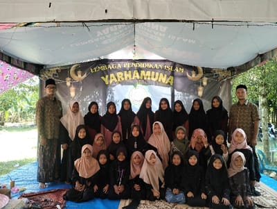An outdoor group portrait in Jambo Dalam, Indonesia, features 29 individuals positioned under a white canopy tent. Two adult males stand on either side of the group. The male on the left wears a patterned shirt, a sarong, and a peci, while the male on the right wears a patterned shirt and a peci. The remaining 27 individuals are females, ranging from young children to young adults, all wearing hijabs and modest clothing, predominantly dark colors. They are arranged in three rows: two standing rows in the back and a seated row of younger girls in the front, all facing the camera. One girl in the seated row is partially covering her face with her hands. Behind the group is a dark banner displaying "LEMBAGA PENDIDIKAN ISLAM YARHAMUNA" in yellow and white text, accompanied by crescent moon and lantern motifs. The ground beneath the tent is covered with a blue tarpaulin, upon which two patterned prayer rugs and miscellaneous items, including a pink bowl and cups, are visible in the left foreground. Beyond the tent, green foliage, trees, and natural ground are visible under bright daylight.