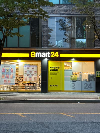 An exterior, eye-level, medium shot captures an E-mart 24 convenience store at dusk on a city street. The store's facade is illuminated by bright yellow signage reading "e-mart24" in large, bold lettering. Below the signage, the storefront features large glass windows offering a view into the well-lit interior. Promotional posters are displayed on the windows. One poster on the left, in Korean and English, advertises "No Brand" products with various item prices listed. A second poster, also in Korean, highlights "No Brand" products and encourages customers to visit. On the right side of the store, large numbers "24" are displayed, part of the store's branding. The store is situated on a paved street with yellow lane markings. Trees with green foliage are visible on either side of the building, their branches partially obscuring the upper floors. The overall impression is of a typical urban convenience store environment. The image was taken in Jeonju-si, South Korea.