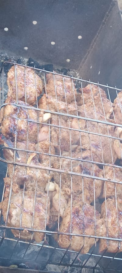 The image shows a close-up view of meat grilling on a barbecue rack. The meat appears to be thinly sliced and seasoned, with visible spices. It is arranged in multiple layers on a metal grill basket, which is placed over hot coals. Smoke is rising from the grill, indicating the cooking process is underway. The background suggests an outdoor setting, with the dark, textured surface of a grill pit visible to the left and bottom of the image. The lighting suggests it is daytime. The scene is devoid of people and text. The focus is entirely on the food being prepared, highlighting the texture and browning of the meat as it cooks.