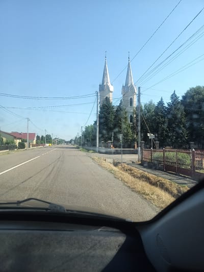 This media file captures a view from a car driving down a road in Siurte, Ukraine. The scene is bathed in bright sunlight, suggesting a clear, warm day, likely midday. The sky is a vibrant, cloudless blue.

The road itself is paved and appears to be a main thoroughfare in a town or village. White lines mark the lanes. On the left side of the road, there are houses with red-tiled roofs, fences, and some greenery. On the right side, a prominent feature is a large church with two identical spires. The church is made of light-colored stone and has gothic architectural elements, including pointed windows and spires topped with crosses. Adjacent to the church, a decorative metal fence encloses the property. Tall, dark green evergreen trees surround the church. Overhead, numerous power lines crisscross the sky, supported by utility poles.

Further down the road, other vehicles are visible, indicating ongoing traffic. A street sign is present on the right side, though the text is partially obscured and difficult to read clearly. A pedestrian crossing with a "zebra" marking is visible near the church. The overall atmosphere is one of a peaceful, rural setting on a bright day. The scene is static, with the only implied activity being the car's movement through the area.