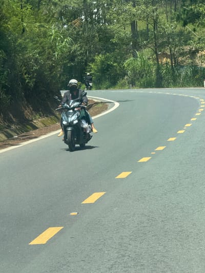 The image shows a person riding a dark-colored scooter on a paved road that curves to the right. The rider is wearing a dark jacket, a light-colored helmet with a pattern, and a dark mask covering their face. They are also wearing yellow footwear. The scooter is a modern design with prominent headlights. Yellow dashed lines are painted on the road to mark the lanes. The road is bordered by a grassy embankment on the left, covered with lush green vegetation, including trees and tall grass. On the right side of the road, the embankment is also vegetated with trees. In the background, more trees and greenery are visible, suggesting a rural or mountainous setting. The sky is bright and appears to be a sunny day. The location is Thôn 6, Vietnam, and the scene depicts a person traveling on a road in a verdant, possibly hilly, area.