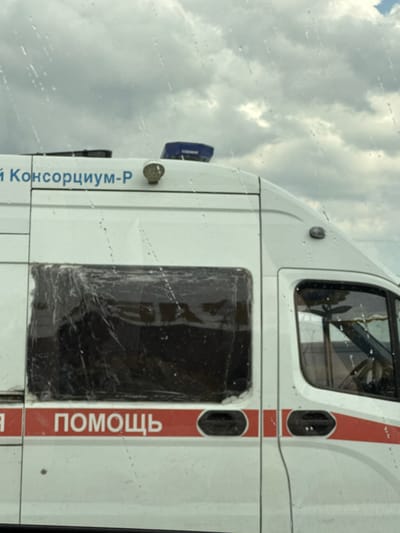 A white ambulance with a red stripe is parked on a street. The ambulance has a black window and some text on its side. The text on the side of the ambulance is in Russian and reads "ПОМОЩЬ" (help) and "Й КОНСОРЦИУМ-Р" (Y Consortium-R). The sky is cloudy, suggesting it may be daytime. The image is taken from a slightly low angle, looking up at the vehicle. There are water droplets on what appears to be a window, possibly indicating rain or a dirty windshield. The overall scene suggests a typical street view of an emergency vehicle in Russia.