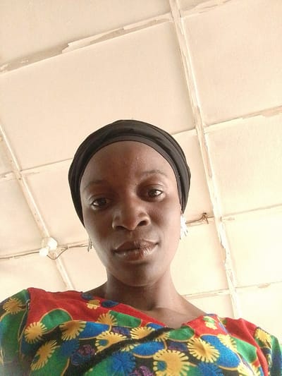 A woman with dark skin and a black head covering is looking down at the camera. She is wearing a brightly colored, floral-patterned top with red, green, yellow, and blue colors. She has small earrings. The background is a stained and aged ceiling with a grid pattern, suggesting an indoor space. There is a light fixture with exposed bulbs visible in the upper-left corner. The location is Barnawa, Nigeria.