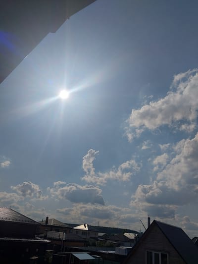 A daytime, low-angle, full shot captures a sunlit sky filled with scattered, puffy white clouds over a residential area. A portion of a dark, angled overhang is visible in the top left corner of the frame, partially obscuring the bright sun which is positioned in the upper left quadrant of the sky and emits prominent light rays. Below the sky, the rooftops of several houses are visible, with varying architectural styles and roof materials, including dark-tiled and lighter-colored pitched roofs. Some of the houses have chimneys. In the background, a low tree line is visible, with power lines and transmission towers interspersed. The scene appears to be a suburban or rural setting in Nizhneye Myachkovo, Russia, characterized by residential buildings and open sky. There are no people or discernible actions depicted in the image. The overall impression is one of a bright, sunny day in a developed area.