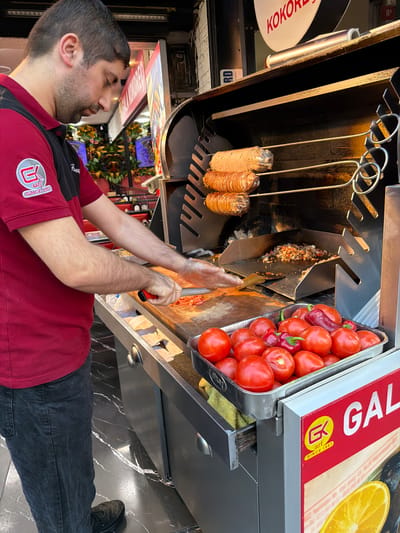 A man in a red and black polo shirt with a circular logo is preparing food in front of a rotating spit of meat, possibly kokoreç, at a street food vendor. He is wearing a red polo shirt with a black panel on the sleeves and shoulder. The logo on his left breast is yellow with a red outline and appears to say "GK" with "SINCE 1987" below it. He is wearing dark denim jeans and black shoes.
The vendor has a large metal grill with the meat rotating on skewers above a hot plate where chopped ingredients are being cooked. In the foreground, a metal tray is filled with ripe red tomatoes and a few red peppers. A cutting board with chopped tomatoes and a red-handled knife are visible on the counter. The vendor is holding a spatula in his right hand, appearing to mix the ingredients on the hot plate.
Behind the vendor, there are signs and decorations, including a red and white sign with Turkish lettering that says "KOKOREÇ". The flooring appears to be black marble with white veins. To the right of the vendor, a red and white sign with "GAL" in large white letters is visible, suggesting the name of the establishment. The overall scene depicts a busy street food environment in the Üsküdar district of Istanbul, Turkey, where this popular dish is commonly served.