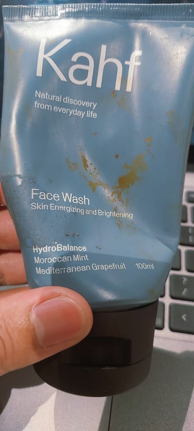 A pale blue tube of Kahf Face Wash with brown and yellow stains is held in a person's hand. The text on the tube reads "Kahf," "Natural discovery from everyday life," "Face Wash," "Skin Energizing and Brightening," "HydroBalance," "Moroccan Mint," "Mediterranean Grapefruit," and "100ml." The background appears to be a laptop keyboard. The image is likely taken in Pagedangan, Indonesia, given the context provided.