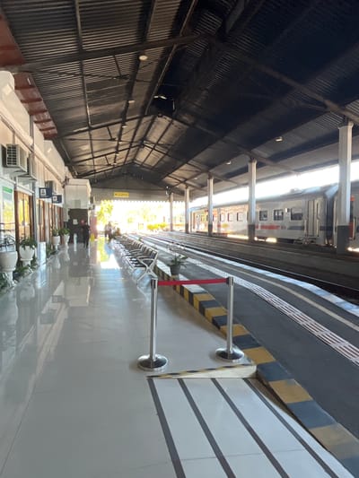 The image shows an outdoor view of a train station platform in Desa Tambakreja, Indonesia. The platform is covered by a dark, corrugated metal roof supported by white pillars. To the left, a white building has several windows and air conditioning units mounted on the exterior. Potted plants are placed along the walkway. The walkway itself is paved with light-colored tiles with reflective surfaces. Several rows of empty white chairs are lined up on the platform. A red rope barrier with stanchions is positioned along the edge of the platform. The train tracks are visible to the right, with a silver train parked alongside. Yellow and black checkered patterns mark the edge of the platform. In the distance, there are glimpses of trees and sunlight. Two people are visible in the background, one walking and others sitting.
