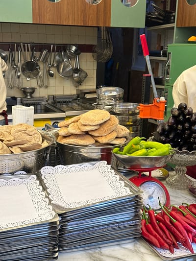 The image depicts a kitchen scene, likely in Cairo, Egypt, given the types of food and kitchenware. The items are arranged on what appears to be a marble countertop.
Objects include:
* **Food:**
* Red chili peppers arranged on a silver tray next to a red scale.
* Green peppers in a silver bowl.
* Pita bread in multiple silver bowls.
* Grapes.
* **Kitchenware:**
* Various stainless steel cooking utensils, including whisks, pots, and ladles, hanging on a rack.
* A manual press, possibly for citrus or other foods.
* Stacks of silver trays with white doilies on top.
* Silver bowls of various sizes.
* **Decor:**
* A decorative glass bowl.
The scene is a mix of ingredients and tools commonly used in a kitchen environment. The focus is on fresh produce and baked goods ready for use, potentially in a restaurant or food preparation setting.
The environment consists of tiled walls and wooden shelves, suggesting a commercial kitchen or food service establishment. There is no presence of people, action or interaction.