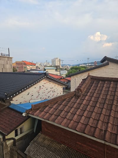An overhead, wide-angle view captures a dense urban landscape with traditional Korean roofs and modern buildings under a partly cloudy sky. The foreground is dominated by the textured, reddish-brown tiles of a pitched roof, with a section of grey, corrugated metal sheeting visible below and to the right. Partially obscured behind this is another roof with dark, rectangular tiles.
Further back, several buildings feature lighter-colored, brick-like facades with decorative trim along the rooflines, reminiscent of traditional Korean architecture, some with bright blue sheeting. A building with an orange-tiled roof is visible to the left of these.
In the mid-ground and background, the urban environment expands to reveal taller, modern buildings and apartment complexes. A prominent, lighter-colored multi-story building with visible windows stands out. Distant, hazy mountains form the backdrop. Overhead, power lines crisscross the sky, and a single, large cumulus cloud is illuminated by the sun on the right side of the frame. The overall scene depicts a transitional urban area, blending traditional and contemporary architectural styles in Jincheon-eup, South Korea. No people or active events are depicted.