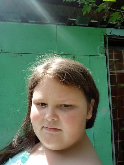 The image, taken in Pavlovsky Posad, Russia, shows a close-up, waist-up portrait of a young girl. She has fair skin, brown hair, and is looking slightly downwards and to the right. Her cheeks are flushed pink. She is wearing a light blue top with a decorative element on the shoulder. The background consists of a weathered, light green corrugated metal wall. Above the wall, the eaves of a structure are visible, with some greenery and small flowers hanging down. A partial view of a dark-framed window or door with a grid pattern is present on the right side of the image. The lighting suggests it is daytime, with sunlight casting highlights on the girl's hair and face, and creating a lens flare effect. The environment appears to be outdoors, possibly in a backyard or near an outbuilding.
