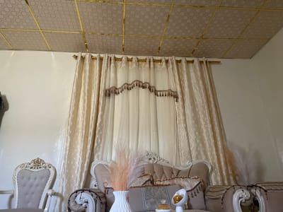 This is a medium shot, eye-level, interior photograph of a room, likely a living room or lounge area, in Hargeisa, Somaliland. The room features ornate, light-colored furniture and elaborate window treatments. The ceiling is paneled with a geometric pattern and accented with gold trim. The walls are painted a neutral color, and a gold curtain rod holds heavy, cream-colored curtains adorned with a decorative valance. In the foreground, a portion of a sofa is visible, upholstered in a light brown fabric with tufted detailing and decorative trim. On the sofa, there is a decorative pillow with a detailed pattern and fringe, and a white vase holding light-colored, feathery floral arrangements. To the left of the sofa is an armchair with a carved, ornate white frame and a light gray tufted seat and back. The overall impression is one of luxurious and decorative interior design. There are no people visible in the image.