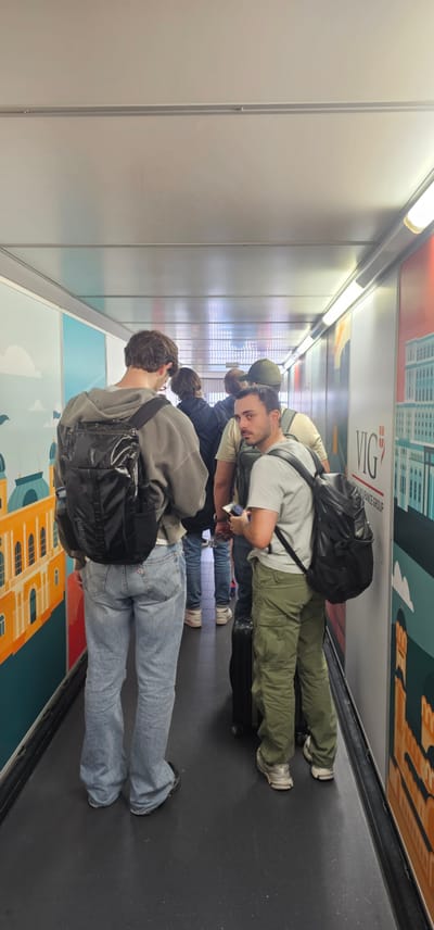 In Fischamend, Austria, individuals are standing in a line within a covered walkway, likely an airport jet bridge or a similar transit area. The walkway has a metallic ceiling with fluorescent lighting. The walls are decorated with large, colorful murals depicting architectural elements and cityscapes, one of which displays the text "VIG INSURANCE GROUP".
The foremost subjects are two men. The man on the left is facing away from the camera, wearing a grey hoodie, light blue jeans, and a black backpack. The man to his right is looking back at the camera, wearing a light grey t-shirt, green cargo pants, and a black backpack. He is holding a blue object in his hand. Behind them, other individuals are visible, also in a line, some with backpacks and luggage, suggesting travel. The floor of the walkway is dark grey.