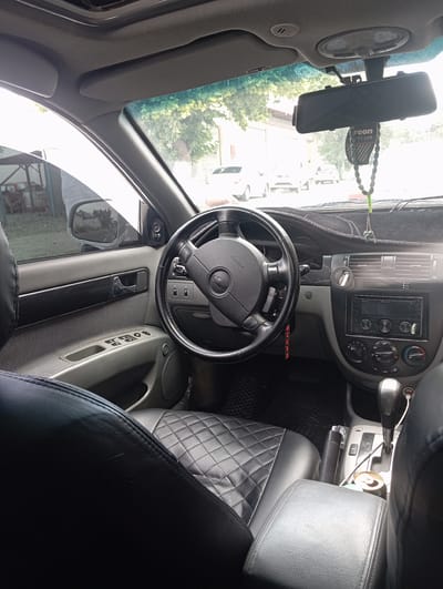 The image shows the interior of a car parked outdoors. The car is a Chevrolet with a black leather interior. The dashboard has a Pioneer stereo system and air conditioning controls. A rearview mirror is mounted on the windshield. Outside the car, trees and other vehicles are visible. The scene appears to be in a residential area.