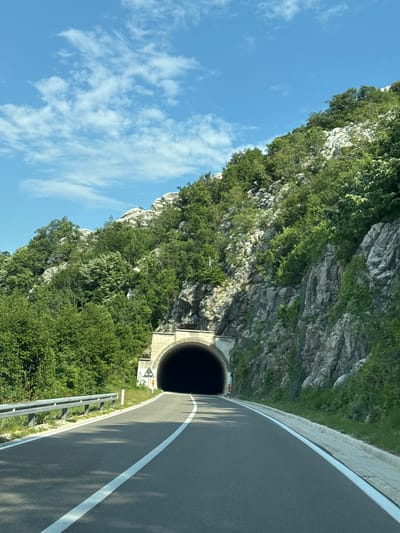 The image shows a view from a vehicle traveling on a paved road towards a tunnel entrance carved into a rocky, vegetated mountainside. The road has white lane markings. A metal guardrail runs along the left side of the road. The sky above is blue with scattered white clouds. The tunnel appears to be a concrete structure with a rounded archway leading into darkness. There are some traffic signs near the tunnel entrance. The surrounding environment is characterized by steep, rocky terrain covered in dense green trees and vegetation. This scene is located in the city of Dragalj, Montenegro.