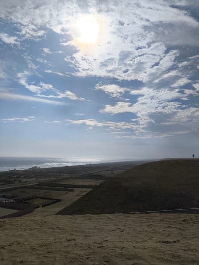 The image shows a coastal view in Huanchaco, Peru, under a sky filled with scattered clouds and bright sunlight. The foreground consists of a sandy dune leading down to a series of cultivated plots and buildings. In the midground, the ocean meets the coastline, and a small bird can be seen flying near the horizon. A small red sign is visible atop a distant hill.