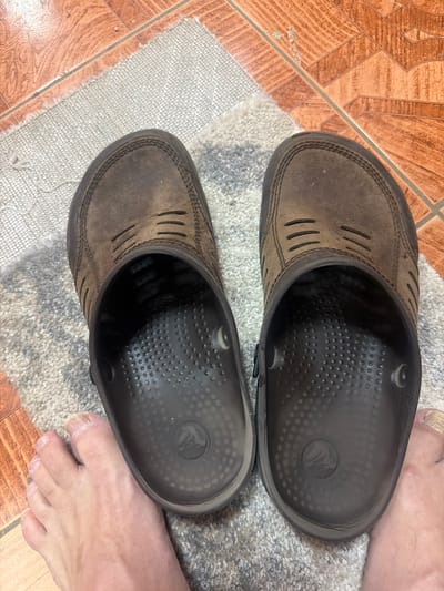 A pair of brown Crocs, appearing worn and slightly distressed, are positioned on a textured gray rug. The rug is placed on a floor with orange tile. The right Croc is closer to the viewer and partially obscures a bare foot, revealing toes with visible nails. The left Croc is positioned next to it, with its interior also visible, showing a textured footbed. A glimpse of another bare foot, similar to the first, is visible to the left of the left Croc. The scene is indoors, with no indication of a specific location in Maloyaroslavets, Russia.