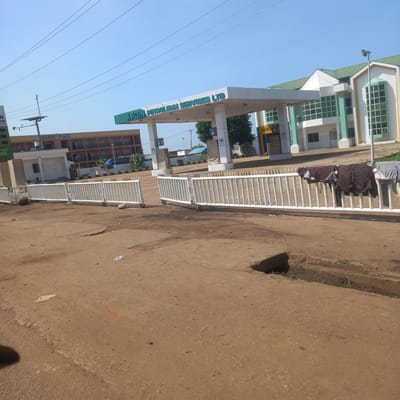 The image depicts a street scene in Hayin Banki, Nigeria. In the foreground, a dirt road with visible tire tracks and debris leads towards a white metal barricade. The barricade appears to be a portable gate or fence on wheels, with some clothing hanging on it. Beyond the barricade, a modern building with a green roof and white and light green facade stands prominently. A covered structure, likely a gas station canopy with signage, is visible in front of the building, bearing the text "AFDIN PETROLEUM SERVICES LTD". Several gas pumps are partially visible under the canopy. To the left, a multi-story building labeled "IDAMBU PLAZA" is visible. The sky is clear and blue. Power lines crisscross the upper portion of the image. The overall environment appears to be a commercial area. There are no people clearly visible in the frame.