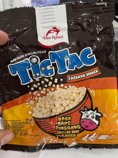 The image shows a snack package labeled "Tic Tac" cassava snack, grilled beef flavor, produced by PT Dua Kelinci Food Industry. The package features a cartoon cow illustration and advertises "Makanan Ringan Pilus." The net weight is 14g. The package also displays production information like the BPOM RI MD number and the manufacturer's address in Pati, Indonesia. It includes information in both Indonesian and Arabic languages. The bag has a brown and orange color scheme.