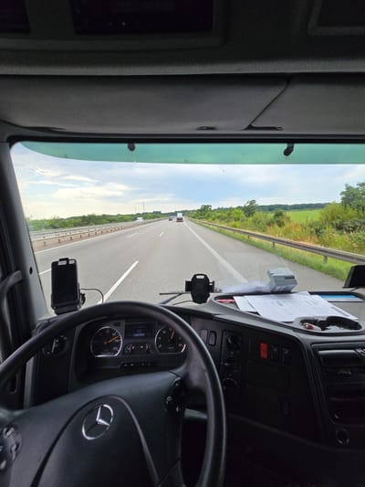 The image is taken from the driver's perspective inside a truck, facing forward. The scene is a highway in Vockerode, Germany. The road stretches ahead into the distance, with white lane markings and a metal guardrail on the right side. Green fields and trees line the highway. The sky is partly cloudy.

Inside the truck's cabin, the steering wheel is visible on the right. The dashboard includes various instruments and controls, a smartphone mounted on a holder, and some papers and a small printer. The time of day appears to be daytime, given the brightness and clear visibility. The weather seems pleasant, with the sky suggesting it's not raining. The overall impression is of a truck in motion on a highway, likely during a journey. There is no visible text that can be read clearly.