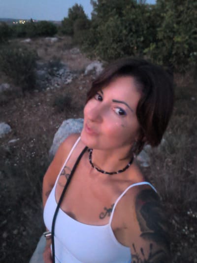 The image displays a female subject in an outdoor, low-light environment identified as Shaalvim, Israel. The woman, appearing in her 20s or 30s, occupies the foreground, looking directly towards the viewer with a slight upward gaze. She has short, dark hair and dark eyes accentuated with blue eyeliner, and small, reflective elements are visible on her left cheekbone. She is wearing a white spaghetti-strap tank top and a dark beaded necklace. Tattoos are present on her right upper arm, left upper arm, and chest area (a stylized symbol). Her right hand, adorned with a silver ring on the middle finger, is visible in the lower left portion of the frame, possibly holding the camera or phone used for the photograph. The background consists of dry, uncultivated terrain with sparse scrub vegetation and scattered rocks. In the far distance, visible under a twilight or nighttime sky, are blurred lights consistent with a distant settlement or city.