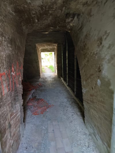 The image depicts a dimly lit, arched stone corridor in Modena, Italy. The walls are constructed of aged brick and rough stone, with some appearing to be partially rendered or plastered. There are areas of visible graffiti in red paint on the left-hand wall. The floor is paved with stones or bricks.
The corridor opens into a brighter area with a path leading outdoors, suggesting an exit or entrance. On the right side of the corridor, there are barred enclosures, reminiscent of windows or cell openings, made of metal grating. Orange construction netting or mesh is piled on the ground near the entrance of the corridor on the left. The overall impression is of an old, possibly abandoned or under-renovation, structure.