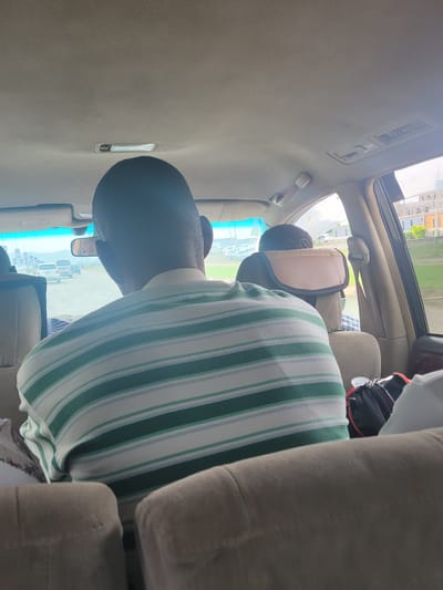 A person is sitting in a vehicle, possibly a car, with a green and white striped shirt. The vehicle appears to have a sunroof. The location shows an urban area in the city of Jiru, Nigeria, discernible through the car window, with buildings and other vehicles visible. The interior of the vehicle is primarily beige.