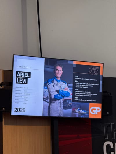A TV screen displays information for "Team GP Elite" driver Ariel Levi. Levi is classified overall, is Israeli, and was born in Tel Aviv on June 13, 2000. He is 171 cm tall and weighs 69 kg. Highlights include 2nd place in the 2025 Porsche Sprint Challenge Southern Europe, 2024 Porsche Sprint Challenge Southern Europe Champion, and 2023 5th place in the Porsche Carrera Cup Benelux. A photo of Levi in racing gear is shown. A "GP" logo is visible.