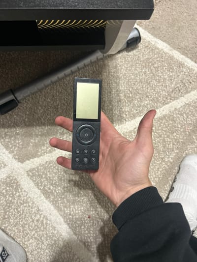 A person's hand holds a black remote control with a blank screen and several buttons, including a circular navigation pad with a power symbol. The hand is wearing a black sweatshirt. The remote is resting on a beige carpet with a subtle geometric pattern. In the background, a portion of a dark furniture item with gold decorative trim is visible, as well as a gray tubular object. The scene is indoors. There is no indication of the city of Ablanitsa, Bulgaria.