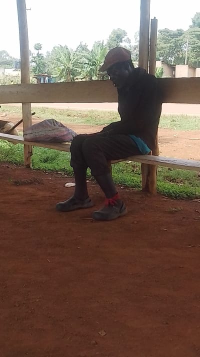 A single adult male is seated on a wooden bench in Siaya Township ward, Kenya. He wears a reddish-brown baseball cap, a dark long-sleeved top, dark trousers, and dark closed-toe shoes worn over dark socks. A red fabric band is tied around his right ankle, above the shoe. His head is tilted downwards. A light blue garment is visible at his lower back, between him and the bench. To his left, a lumpy, grayish bag rests on the bench. The immediate ground beneath is reddish-brown earth. Behind the bench, green grass transitions to a lighter-colored strip of ground, followed by dense vegetation, including numerous banana trees. Faint outlines of distant structures are visible in the far background under a bright, overcast sky.