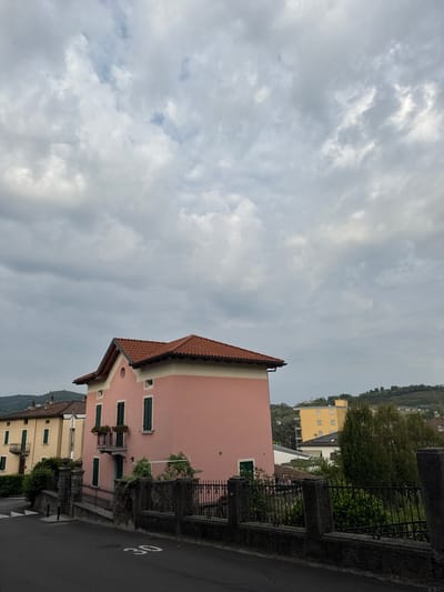 This is a medium-shot, exterior, eye-level, vertical, daytime photograph of a street scene in Balerna, Switzerland. The sky is heavily overcast with grey clouds. In the foreground, a paved road with a speed limit sign reading "30" is visible. A black metal fence runs along the side of the road, separating it from a property. Behind the fence, a pink building with a terracotta tile roof stands prominently. It has green shutters on its windows and a small balcony with planters. To the left of the pink building, a yellow building with similar architectural features is partially visible. In the background, there is a modern yellow building and a verdant hillside with trees and terraced vegetation. The overall impression is of a quiet, residential street in a town with hilly surroundings. There are no people or animals visible in the image.