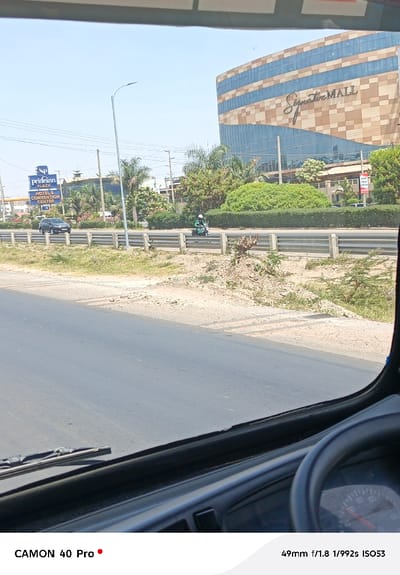 From inside a vehicle, a view of a road. A motorcyclist in green is on the other side of a median. Further away is a shopping complex labeled “Signature Mall.” A PrideInn Plaza Hotel and Convention Centre sign is visible. The sky is light blue, and the sun appears bright. The speedometer and parts of the dashboard are also visible in the lower right corner of the frame.