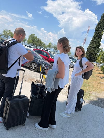 In Mojanovići, Montenegro, three individuals stand outdoors with luggage. The scene is a parking lot, with cars visible in the background. A tall tower with red and white markings stands in the distance, suggesting a possible airport or communication hub. The sky is a vibrant blue with scattered white clouds, indicating clear weather. The sunlight suggests it is daytime, likely late morning or early afternoon.

Two women and one man are present, all appearing to be travelers. The woman closest to the camera is wearing a white t-shirt and light-colored pants, with a black backpack slung over her shoulder. She is looking down and to the left, her expression unreadable due to her downward gaze. The second woman, in the middle, is wearing a white short-sleeved top and black pants. She has her hair pulled back in a bun and is looking to her left. A grey sweater is draped over her arm, and she is holding a dark bottle. The man in the foreground is seen from the back, wearing a white t-shirt and black pants. He has a backpack on and is looking to his right, seemingly towards the other individuals.

Several pieces of luggage are visible: two black suitcases are placed on the ground, one near the middle woman and another further to the right. The man also has a black backpack. One of the suitcases has a tag with visible text, including "TGD". The overall mood seems to be one of travel, with the individuals likely preparing for or having just arrived from a journey.