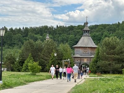 The image captures a daytime scene in Vzglyadnevo, Russia. A concrete walkway leads towards a tall, ornate wooden gate with a prominent spire topped with a cross. A smaller wooden tower is visible further back among the trees. A group of approximately ten people of varying ages walks along the walkway in the direction of the gate. The people are dressed in casual summer clothing. The environment consists of a grassy area on either side of the walkway, with trees and shrubbery in the background, which form the backdrop of the gate. Two black lampposts flank the walkway on either side of the frame. The sky is overcast with some patches of blue. A sign in Russian is visible near the gate.