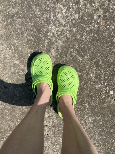 In an outdoor scene, a pair of neon green clogs are worn by a person standing on a cracked concrete surface. The shoes are the central focus, positioned at the top of the frame, with the wearer's legs extending downwards. The concrete ground shows signs of wear and tear, with visible imperfections and variations in color. Sunlight casts a shadow from the left shoe, indicating a bright day. The image is an overhead shot, offering a clear view of the footwear and the ground. The location is not specified in the image itself.