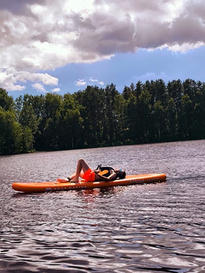 A person is lying on their back on an orange paddleboard in a body of water. The paddleboard has text that reads "AQUA MARINA FUSION 10'10" X 32"". The person is wearing orange shorts and a life vest. They are wearing sunglasses. The water is rippled and reflects the sky and trees. In the background, there is a dense line of green trees along the shore of the body of water. Above the trees, there are white and grey clouds in a blue sky. The location is identified as Vecumnieki, Latvia.