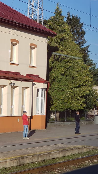 The image shows a train station platform in Spišská Nová Ves, Slovakia. Two men are standing on the platform. The man on the left is wearing a red polo shirt, blue jeans, and a blue baseball cap. He is standing with his arms crossed and looking to the right. The man on the right is wearing a dark blue sweater and dark blue jeans. He is standing with his hands in his pockets and looking to the left. In the background, there is a building with a red roof and a large green tree. There are also train tracks in the foreground. The sky is clear and blue.