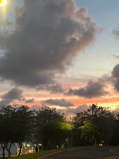The image captures a sky at dusk or dawn in Le Gosier, France, evidenced by the tropical flora and lighting fixtures. Dominating the view are dark, billowing storm clouds tinged with pink and orange light from the setting or rising sun. The light creates a gradient of color on the horizon. To the right, trees line a path or road, their dark silhouettes interspersed with sporadic lights along the path. Faint human figures can be discerned by the waterside in front of one or two light posts. The perspective is slightly tilted, which may be from the orientation of the photograph.