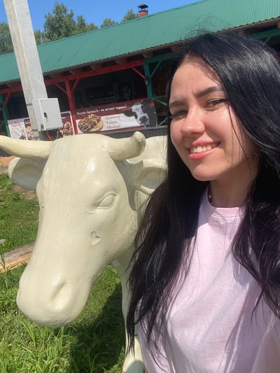 A young woman with long, dark hair and a light pink t-shirt smiles at the camera, taking a selfie. She is standing next to a cream-colored statue of a cow. In the background, there is a building with a green roof and red trim, which appears to be a restaurant or cafe, possibly with outdoor seating. Signs are visible on the building, one featuring an image of kebabs and another with text in Cyrillic. The setting is outdoors, with grass and trees visible. This location is in the city of Verkhnii Liuk, Russia.