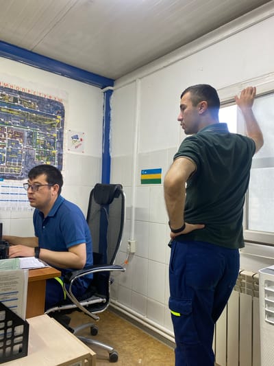 The image depicts an indoor scene, likely an office or control room, with two men. The man on the left is seated at a desk, wearing a blue polo shirt and glasses, and appears to be working on a computer. He has a radio on his desk. The man on the right is standing, facing away from the camera and towards a window. He is wearing a dark green polo shirt and blue cargo pants with a yellow reflective strip. He has his left hand on the window frame and his right hand on his hip. The environment includes a map on the wall, a chair, a radiator, and some documents. There is a small flag on the wall with horizontal stripes of blue, yellow, and green, possibly indicating the flag of Gabon. The overall impression is of a functional, possibly industrial, workspace. The location is stated to be in the city of Yukhta, Russia, though there are no visual cues within the image to confirm this specific location.