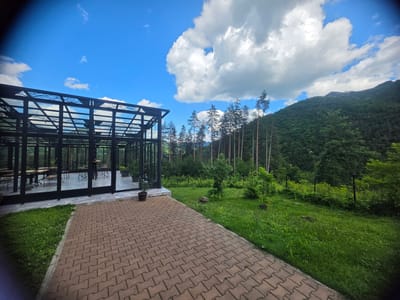 A wide-angle, eye-level shot captures a modern greenhouse structure with a black metal frame and glass walls, situated in a lush, green, mountainous environment. The greenhouse contains tables and chairs, suggesting it might be a restaurant or event space. A paved walkway made of interlocking brick leads from the foreground towards the greenhouse. To the right of the walkway is a well-maintained lawn dotted with young trees and a black metal fence running along the edge of the property. The background features a dense forest covering a steep mountainside, with tall, thin pine trees in the middle ground. The sky above is a vibrant blue, with fluffy white clouds and a hazy sun. The scene is peaceful and idyllic, evoking a sense of tranquility. While the image does not explicitly depict any people or specific actions, the presence of the greenhouse and its furnishings implies human activity and purpose. The location appears to be in or near Teteven, Bulgaria, a region known for its natural beauty and mountainous terrain.