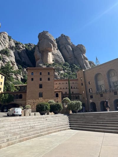 The image shows a view of Montserrat, a mountain range located near Monistrol de Montserrat, Spain. In the foreground, there is a large, wide staircase made of light-colored stone. To the right, a man wearing a black t-shirt and black shorts is walking up the stairs. Behind the stairs, there are several buildings, including a large, tan-colored structure with arched entrances and decorative reliefs on its facade. To the left of this building, a multi-story building with a red-tiled roof and brick facade is visible. Trees and potted plants are situated in front of the buildings. In the background, the rugged, rocky peaks of Montserrat rise steeply against a clear blue sky. A white van with a logo on its side is parked to the left of the stairs. The sun is shining brightly, casting shadows on the scene.