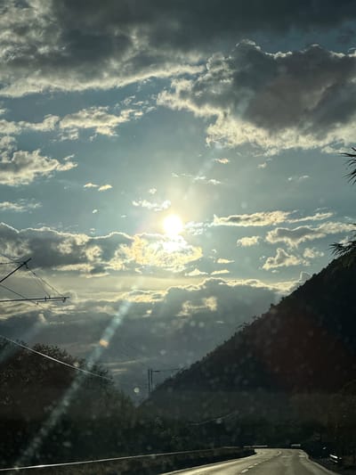 The image captures an expansive sky and a natural landscape in Vranjina, Montenegro, likely seen through a vehicle window. The sky dominates the upper and left portions of the frame, filled with a dynamic arrangement of clouds. The sun, a brilliant disc, is positioned in the upper-left quadrant, partially veiled by clouds. Its intense light illuminates the edges of the clouds with golden hues and produces prominent lens flares and sunrays that stretch across the sky. Some clouds are dense and dark, contrasting with the brighter, elongated formations that glow from the sun's light.
Towards the right side of the image, a dark, tree-covered hillside or mountain rises, bordering a paved road with a clear white line visible at the bottom-right. In the lower-left to mid-left, utility poles and electrical wires crisscross the frame. Dark foliage, possibly from a tree close to the camera, is visible along the very top-left edge. Numerous circular and streak-like reflections and small specks, indicative of dust or water on a window, are present throughout the image. The dramatic lighting, with the sun low in the sky, strongly suggests either sunrise or sunset, creating a powerful and atmospheric scene. No people or visible text are present.