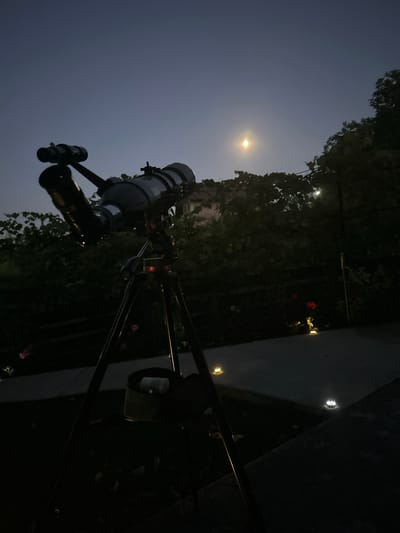 The image shows a telescope set up outdoors at night. The telescope is a dark gray cylinder mounted on a tripod. It is pointed upwards towards a bright full moon. The background consists of dark trees and a clear, dark blue sky. There are also some small, glowing lights visible in the distance, possibly streetlights or garden lights. The overall scene suggests an evening of stargazing. The image is oriented sideways.