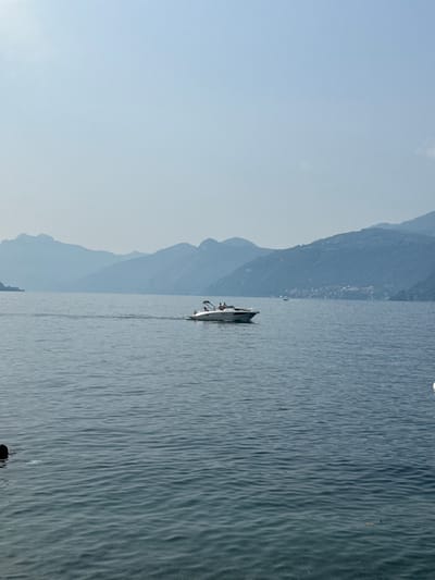 A wide shot shows a body of water, likely Lake Como, with mountains in the background. A motorboat with at least two occupants is moving across the water. The boat is white and appears to be a speed boat. The mountains are hazy and blue, suggesting distance or light fog. The sky is a pale blue. The water is choppy with small waves. In the foreground, there are some dark shapes in the water that may be buoys or debris. The image is taken from a slightly elevated position, looking out over the water.