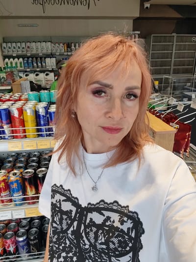 The media file shows a selfie of a woman with pink hair taken in a store, possibly a convenience store or a supermarket. She is wearing a white t-shirt with a black lace-like print on the front. The background is filled with shelves stocked with various products, including rows of canned drinks and bottles of liquids. Some of the visible text on the cans and shelves includes "Red Bull", "Energy Drink", "The Blue Edition", and Russian Cyrillic text which is partially obscured or illegible in the provided crops. The woman is looking directly at the camera with a neutral expression, and her makeup includes eyeshadow and lipstick. The lighting suggests it could be daytime, but the interior setting makes it difficult to determine the exact time of day or weather conditions. The overall impression is that of a casual snapshot taken while shopping.
